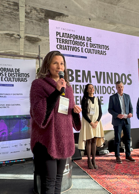 Mulher com suéter vinho fala ao microfone em um palco. Atrás dela, a mesma tela digital exibe a frase “BEM-VINDOS / BIENVENIDOS”, e outras três pessoas (duas mulheres e um homem) observam com atenção. O cenário inclui plantas e um banner institucional do evento.




