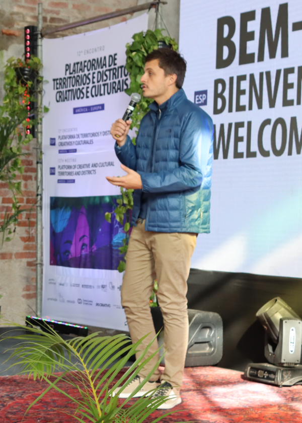 Foto de Pedro Valério, diretor-executivo do Instituto Caldeira, em pé sobre um palco, segurando um microfone enquanto fala ao público. Ele veste uma jaqueta azul acolchoada, calça bege e tênis claros. Ao fundo, há um cartaz do evento com o título "Plataforma de Territórios e Distritos Criativos e Culturais – América-Europa" e a palavra "BEM-VINDO" escrita em vários idiomas. O ambiente tem estilo industrial, com parede de tijolos aparentes e decoração com plantas. A iluminação natural destaca a cena e parte de um tapete oriental cobre o palco.