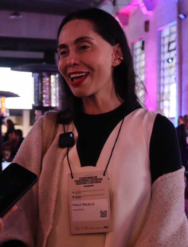 Foto de Paula Trujillo, enquanto é entrevistada. Ela usa um vestido branco sobre blusa preta e crachá que a identifica como “Palestrante”. Ela fala enquanto utiliza um microfone preso à roupa. O ambiente ao fundo está com iluminação roxa e pessoas circulando.

