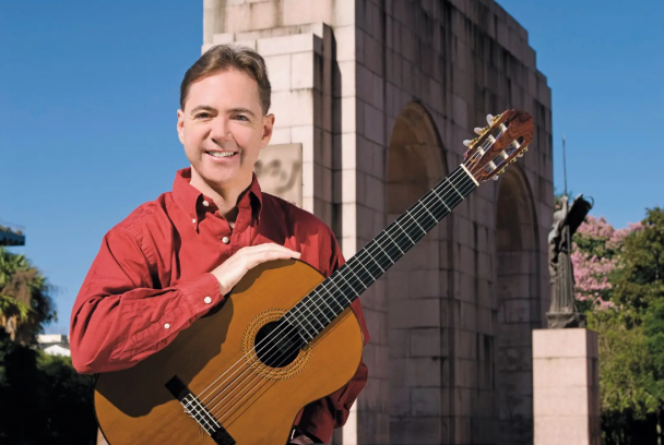 Foto do violonista Daniel Wolff, vestindo uma camisa vermelha e segurando um violão acústico, diante do Monumento ao Expedicionário, no Parque Farroupilha em Porto Alegre. Ao fundo, o céu azul claro contrasta com a vegetação esparsa de árvores