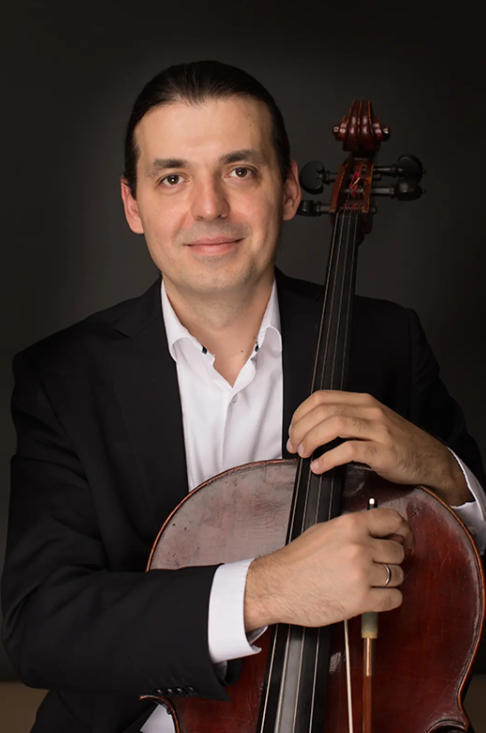 Foto do violoncelista sérvio-americano Viktor Uzur, com traje formal: terno preto e camisa branca. Ele segura um violoncelo contra um fundo escuro. As mãos estão posicionadas no braço e no corpo do instrumento, posando para a foto.