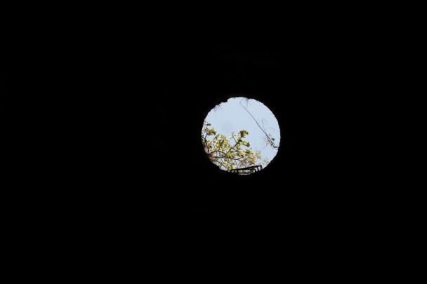 Fotografia escura, onde tudo ao redor está completamente preto, exceto por um círculo no centro da imagem. Esse círculo parece ser uma abertura redonda no teto, como um respiradouro ou claraboia, através da qual se vê o céu azul e parte de uma árvore com folhas verdes iluminadas pela luz natural.