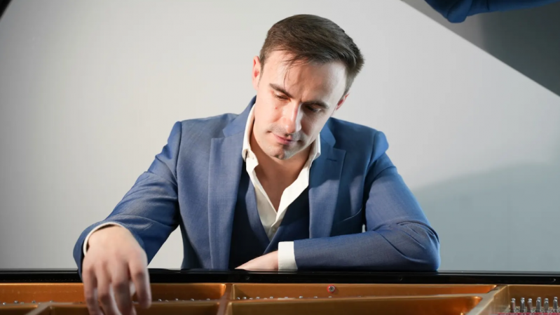 Foto do pianista britânico Connor Heraghty, de terno azul, com camisa branca parcialmente aberta, sentado diante de um piano de cauda, com a tampa levantada. Ele olha atentamente para as cordas e martelos do instrumento, enquanto estende uma das mãos para o interior do piano, sugerindo que está examinando sua mecânica. A iluminação suave destaca sua expressão concentrada e o ambiente tem fundo neutro.