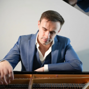 Foto do pianista britânico Connor Heraghty, de terno azul, com camisa branca parcialmente aberta, sentado diante de um piano de cauda, com a tampa levantada. Ele olha atentamente para as cordas e martelos do instrumento, enquanto estende uma das mãos para o interior do piano, sugerindo que está examinando sua mecânica. A iluminação suave destaca sua expressão concentrada e o ambiente tem fundo neutro.
