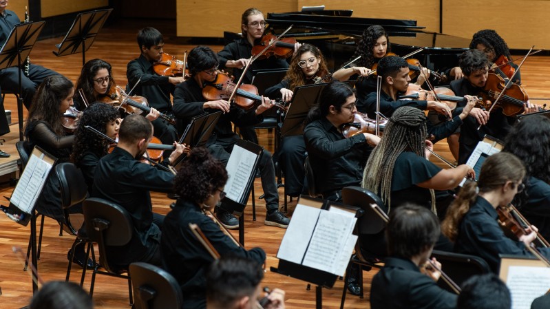 Fotografia de uma orquestra jovem em apresentação. O grupo é formado por diversos músicos sentados em cadeiras dispostas em semicírculo, todos vestindo roupas pretas. A maioria toca instrumentos de corda, como violinos e violas, concentrados na execução da música. Partituras estão apoiadas em estantes à frente de cada músico. Ao fundo, vê-se um piano de cauda e painéis de madeira clara cobrindo as paredes. O ambiente é uma sala de concertos com piso de madeira, iluminada por luzes superiores.
