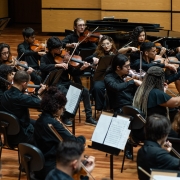 Fotografia de uma orquestra jovem em apresentação. O grupo é formado por diversos músicos sentados em cadeiras dispostas em semicírculo, todos vestindo roupas pretas. A maioria toca instrumentos de corda, como violinos e violas, concentrados na execução da música. Partituras estão apoiadas em estantes à frente de cada músico. Ao fundo, vê-se um piano de cauda e painéis de madeira clara cobrindo as paredes. O ambiente é uma sala de concertos com piso de madeira, iluminada por luzes superiores.