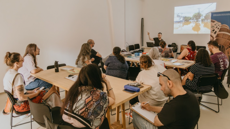 Em uma sala da Casa de Cultura Mario Quintana, com paredes brancas e iluminação natural, um grupo de pessoas está reunido em um ambiente de oficina ou curso. Elas estão sentadas em torno de duas mesas retangulares de madeira clara, voltadas para um homem que apresenta um conteúdo projetado na parede à frente. Ele está em pé ao lado de um notebook e gesticula com a mão levantada enquanto fala. Na projeção, vê-se uma imagem de rua urbana com alagamento. Os participantes, de diferentes idades e estilos, prestam atenção, fazem anotações ou observam o conteúdo. Há materiais diversos sobre as mesas, como livros, cadernos, lápis e garrafas de água. À direita, um banner colorido mostra parte de uma edificação arquitetônica clássica. A atmosfera sugere um encontro educativo e colaborativo.