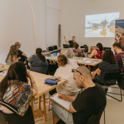 Em uma sala da Casa de Cultura Mario Quintana, com paredes brancas e iluminação natural, um grupo de pessoas está reunido em um ambiente de oficina ou curso. Elas estão sentadas em torno de duas mesas retangulares de madeira clara, voltadas para um homem que apresenta um conteúdo projetado na parede à frente. Ele está em pé ao lado de um notebook e gesticula com a mão levantada enquanto fala. Na projeção, vê-se uma imagem de rua urbana com alagamento. Os participantes, de diferentes idades e estilos, prestam atenção, fazem anotações ou observam o conteúdo. Há materiais diversos sobre as mesas, como livros, cadernos, lápis e garrafas de água. À direita, um banner colorido mostra parte de uma edificação arquitetônica clássica. A atmosfera sugere um encontro educativo e colaborativo.