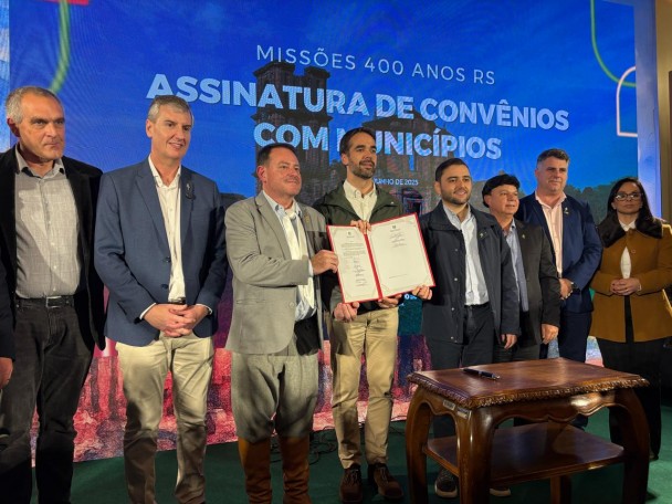 Em um palco, o governador Eduardo Leite segura um documento acompanhado do Voce-governador Gabriel Souza, dos Secretários Ronaldo Santini, do Turismo, Eduardo Loureiro, da Cultura, e prefeitos de municípios da região das missões. 