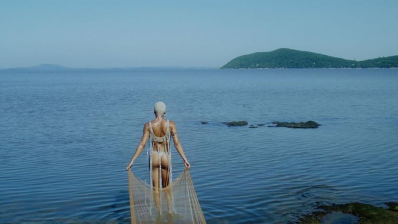 Pessoa de costas entra em um corpo d'água calmo, vestindo um traje claro e translúcido com franjas que lembram uma rede de pesca. A figura central está parcialmente submersa e caminha em direção ao horizonte, onde se avistam ilhas ou morros cobertos por vegetação.