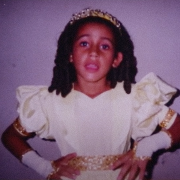 Fotografia antiga de uma criança posando em frente a uma parede lisa. Ela está fantasiada de princesa, usando um vestido amarelo claro com detalhes dourados, mangas bufantes, luvas brancas curtas e uma tiara brilhante.