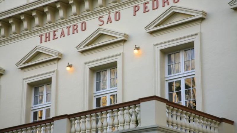 Fachada do Theatro São Pedro, em Porto Alegre, com destaque para a varanda com balaústres e três janelas envidraçadas. Acima delas, vê-se a inscrição "THEATRO SÃO PEDRO" em letras vermelhas. A arquitetura é de estilo neoclássico, com ornamentos como frontões triangulares sobre as janelas e detalhes em relevo. Três luminárias acesas iluminam a fachada de tom bege claro.