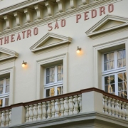 Fachada do Theatro São Pedro, em Porto Alegre, com destaque para a varanda com balaústres e três janelas envidraçadas. Acima delas, vê-se a inscrição "THEATRO SÃO PEDRO" em letras vermelhas. A arquitetura é de estilo neoclássico, com ornamentos como frontões triangulares sobre as janelas e detalhes em relevo. Três luminárias acesas iluminam a fachada de tom bege claro.