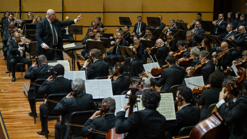 Imagem da Ospa, em apresentação, capturada de um ângulo lateral que evidencia a extensão do grupo. Em primeiro plano, músicos de costas tocam instrumentos de corda, como violoncelos e contrabaixos, com partituras abertas à frente. Ao centro, o maestro Manfredo Schmiedt, um homem usando terno preto e gravata listrada, rege com a mão direita levantada e a esquerda próxima à partitura sobre o pódio. À sua frente, está a seção de cordas com diversos violinistas concentrados. No fundo, são visíveis músicos de sopro e percussão, além de uma fileira de coralistas ou músicos de apoio. O cenário é composto por piso de madeira e painéis acústicos claros, criando um ambiente formal e acústico apropriado.
