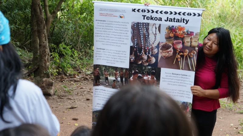 Mulher indígena com cabelos longos e lisos, vestindo blusa vermelha, segura um banner informativo em meio à natureza, com vegetação verde ao fundo. O banner traz o título “Tekoa Jataity” e informações bilíngues (em português e guarani) sobre o artesanato, danças, cantos e alimentação tradicional da comunidade. Também há fotografias de cestos e instrumentos artesanais, além de imagens de membros da comunidade em atividades culturais. 