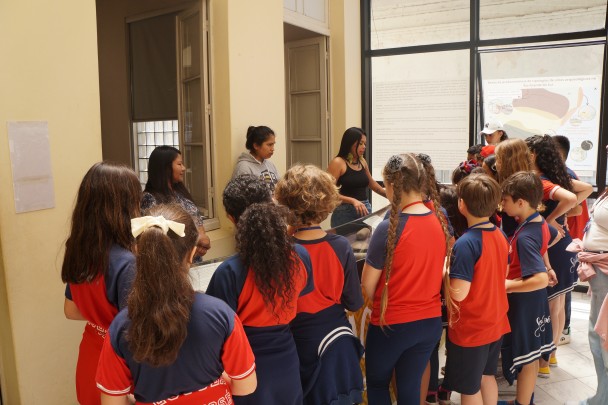 Grupo de crianças usando uniformes nas cores azul e vermelho, observa com atenção uma exposição em ambiente interno, iluminado por luz natural. Elas estão reunidas em frente a uma vitrine de vidro com objetos expostos. Três mulheres indígenas estão posicionadas atrás da vitrine, explicando os elementos apresentados. Uma delas fala enquanto aponta para a vitrine; outra está sorrindo e a terceira observa atenta.