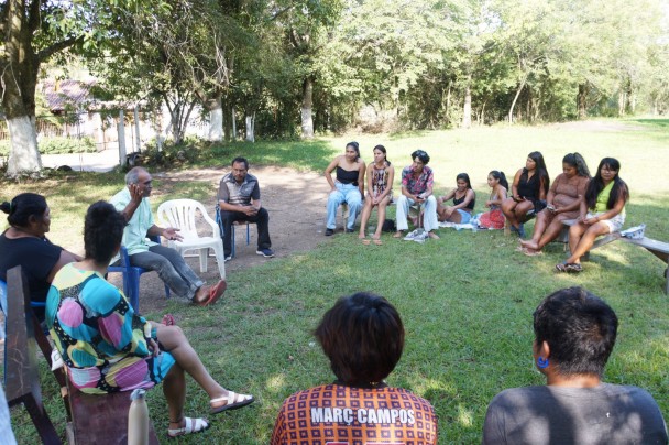 Grupo de pessoas sentadas em círculo ao ar livre, participando de uma roda de conversa sob a sombra de árvores. O ambiente é gramado e cercado por vegetação, com clima informal e acolhedor. Um homem de camisa verde claro gesticula enquanto fala, enquanto os demais escutam com atenção.