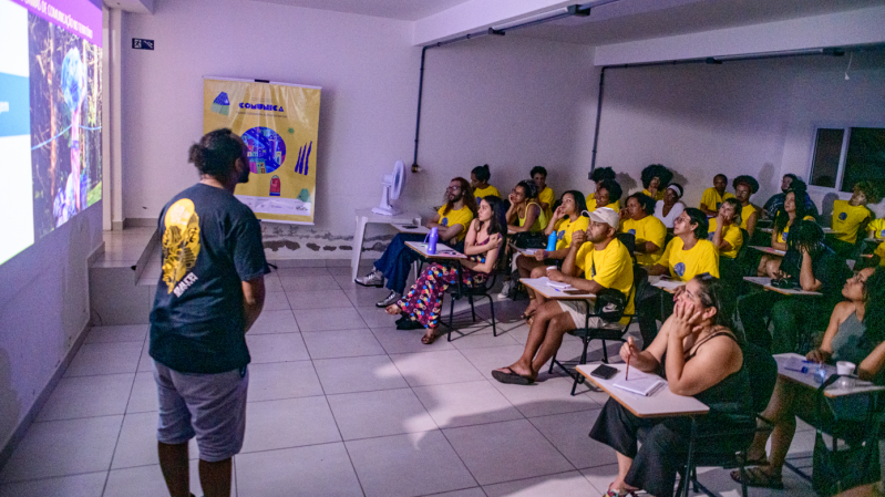 Foto durante uma oficina do projeto Projeto Comunica. É uma sala de aula com iluminação baixa, onde uma pessoa está em pé à frente, apresentando algo projetado na parede para um grupo atento de participantes. A maioria está vestindo camisetas amarelas e sentada em carteiras escolares, fazendo anotações ou observando a apresentação. Ao fundo, há um banner do projeto, escrito “Comunica” e ilustrações coloridas. O ambiente é simples, com paredes brancas e piso de cerâmica clara.