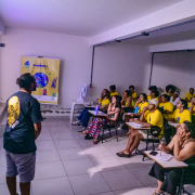 Foto durante uma oficina do projeto Projeto Comunica. É uma sala de aula com iluminação baixa, onde uma pessoa está em pé à frente, apresentando algo projetado na parede para um grupo atento de participantes. A maioria está vestindo camisetas amarelas e sentada em carteiras escolares, fazendo anotações ou observando a apresentação. Ao fundo, há um banner do projeto, escrito “Comunica” e ilustrações coloridas. O ambiente é simples, com paredes brancas e piso de cerâmica clara.