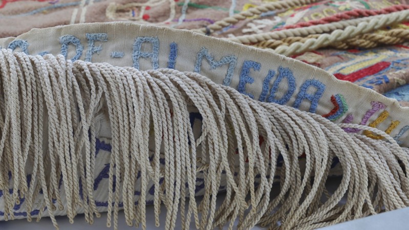 Imagem em close de uma peça têxtil artesanal, composta por bordados coloridos sobre tecido bege e detalhes em relevo com fios grossos e torcidos.
