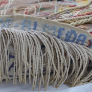 Imagem em close de uma peça têxtil artesanal, composta por bordados coloridos sobre tecido bege e detalhes em relevo com fios grossos e torcidos. 