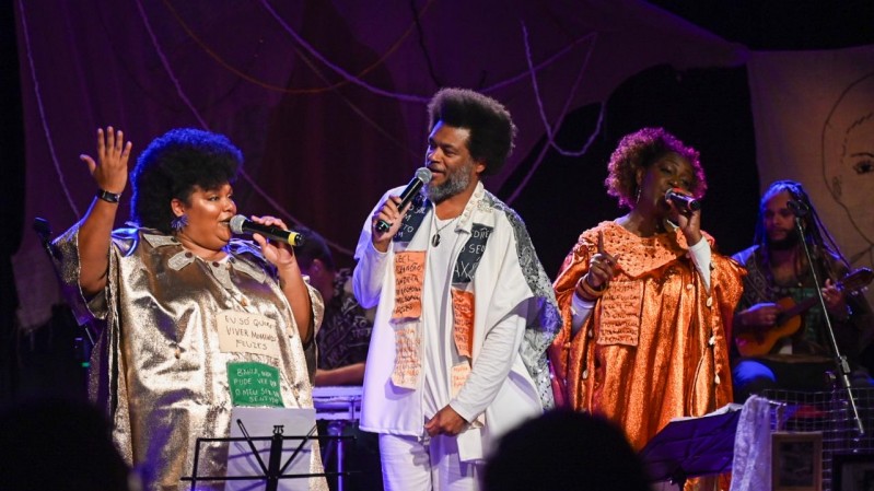 Três cantores negros se apresentam no palco, cada um segurando um microfone e cantando com expressividade. À esquerda, uma mulher usa um vestido dourado brilhante com frases bordadas. Ao centro, um homem veste roupa branca com panos estampados com textos, e à direita, outra mulher usa uma túnica laranja cintilante também decorada com frases escritas. Ao fundo, um músico toca um instrumento de cordas O cenário é escuro, com iluminação focada nos artistas e elementos decorativos ao fundo, como tecidos e desenhos.