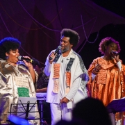 Três cantores negros se apresentam no palco, cada um segurando um microfone e cantando com expressividade. À esquerda, uma mulher usa um vestido dourado brilhante com frases bordadas. Ao centro, um homem veste roupa branca com panos estampados com textos, e à direita, outra mulher usa uma túnica laranja cintilante também decorada com frases escritas. Ao fundo, um músico toca um instrumento de cordas O cenário é escuro, com iluminação focada nos artistas e elementos decorativos ao fundo, como tecidos e desenhos. 