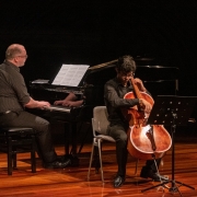 Dois músicos se apresentam em um palco de madeira com iluminação direcionada. À esquerda, um pianista calvo e de óculos, vestido com camisa escura e calça preta, toca um piano de cauda com partituras abertas. À direita, um jovem violoncelista, também trajando roupas escuras, toca seu instrumento sentado, com um estante de partituras à sua frente.