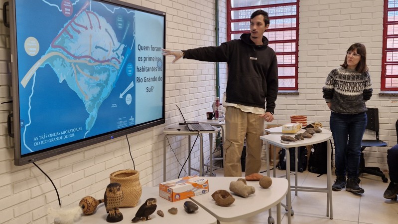 A imagem mostra duas pessoas em sala de aula, uma delas aponta para uma lousa digital com apresentação em que aparece o mapa do Rio Grande do Sul. A frente deles há uma mesa com objetos do acervo museológico enfileiradas. 