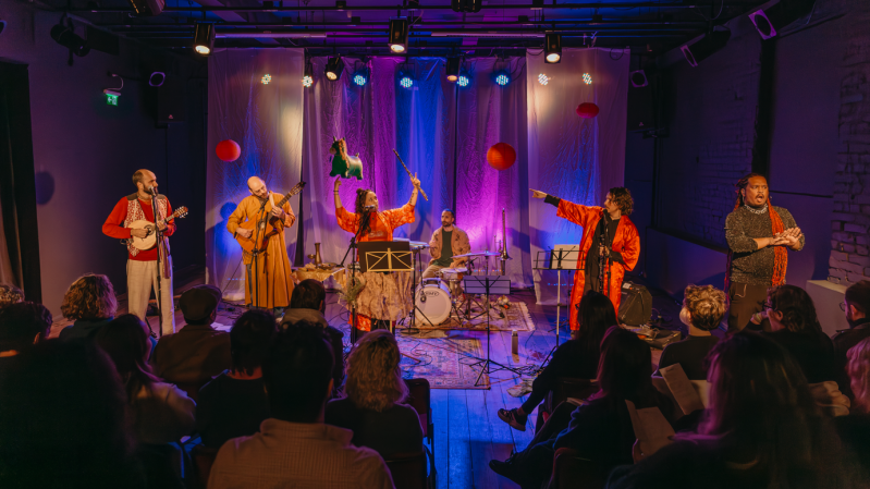 Foto da apresentação “Canções para suportar o fim dos tempos”, mostrando um grupo de artistas se apresenta em no palco do Teatro Carlos Carvalho, iluminado com luzes coloridas, em tons de roxo, azul e âmbar, diante de uma plateia sentada. Ao centro, uma mulher com roupa vermelha levanta um pequeno burro de brinquedo verde e uma vareta com fitas, enquanto canta ao microfone. À sua volta, músicos tocam instrumentos como cavaquinho, violão e bateria. À direita, uma pessoa de casaco preto realiza interpretação em Libras. O cenário é decorado com lanternas vermelhas e cortinas translúcidas, criando um clima festivo e teatral.