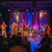 Foto da apresentação “Canções para suportar o fim dos tempos”, mostrando um grupo de artistas se apresenta em no palco do Teatro Carlos Carvalho, iluminado com luzes coloridas, em tons de roxo, azul e âmbar, diante de uma plateia sentada. Ao centro, uma mulher com roupa vermelha levanta um pequeno burro de brinquedo verde e uma vareta com fitas, enquanto canta ao microfone. À sua volta, músicos tocam instrumentos como cavaquinho, violão e bateria. À direita, uma pessoa de casaco preto realiza interpretação em Libras. O cenário é decorado com lanternas vermelhas e cortinas translúcidas, criando um clima festivo e teatral.