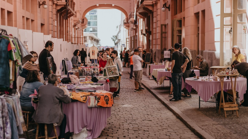 A imagem mostra a "Feira Sem Título", realizada na Travessa Rua dos Cataventos, dentro da Casa de Cultura Mario Quintana. Diversas barracas cobertas com toalhas roxas estão dispostas ao longo do espaço, expondo itens variados como roupas, artesanatos, quadros e produtos naturais. Pessoas circulam, observam os produtos e conversam entre si, criando um ambiente movimentado e acolhedor. Ao fundo, é possível ver um arco que conecta os prédios e emoldura a continuação da rua. A luz do sol ilumina suavemente o local, reforçando a atmosfera agradável da cena.
