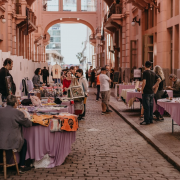 A imagem mostra a "Feira Sem Título", realizada na Travessa Rua dos Cataventos, dentro da Casa de Cultura Mario Quintana. Diversas barracas cobertas com toalhas roxas estão dispostas ao longo do espaço, expondo itens variados como roupas, artesanatos, quadros e produtos naturais. Pessoas circulam, observam os produtos e conversam entre si, criando um ambiente movimentado e acolhedor. Ao fundo, é possível ver um arco que conecta os prédios e emoldura a continuação da rua. A luz do sol ilumina suavemente o local, reforçando a atmosfera agradável da cena.
