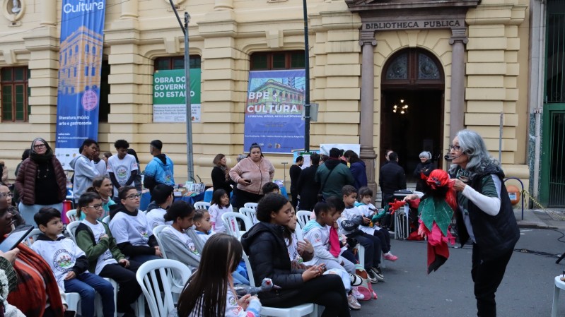 Várias pessoas caminham e observam estandes de livros expostos em frente à Biblioteca Pública do Estado do RS, cuja fachada amarela aparece ao fundo. Grandes banners verticais e horizontais anunciam o evento “BPE + Cultura – Literatura, Música, Arte e Gastronomia”. 