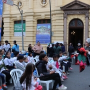 Várias pessoas caminham e observam estandes de livros expostos em frente à Biblioteca Pública do Estado do RS, cuja fachada amarela aparece ao fundo. Grandes banners verticais e horizontais anunciam o evento “BPE + Cultura – Literatura, Música, Arte e Gastronomia”. 