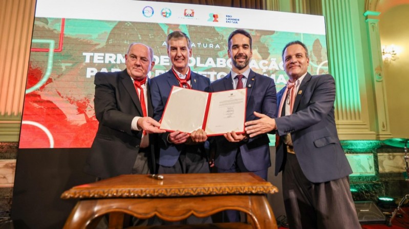 Foto que mostra o secret&aacute;rio da Cultura, Eduardo Loureiro, e o governador do Rio Grande do Sul, Eduardo Leite, acompanhados de dois representantes do Movimento Tradicionalista Ga&uacute;cho. Os quatro posam sorrindo, segurando juntos um documento aberto &agrave; frente, no Sal&atilde;o Negrinho do Pastoreio do Pal&aacute;cio Piratini. Todos est&atilde;o vestidos formalmente, sendo que tr&ecirc;s usam indument&aacute;ria tradicional ga&uacute;cha. &Agrave; frente deles, h&aacute; uma mesa de madeira trabalhada. 
