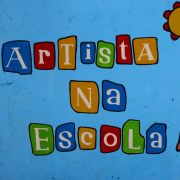 Mural colorido com fundo azul claro onde se lê a frase “Artista na Escola” escrita com letras brancas dentro de formas coloridas que lembram blocos infantis. O mural foi pintado por alunos da Escola Estadual de Ensino Médio Antão de Faria, no bairro Bom Jesus, em Porto Alegre. As letras estão distribuídas em três linhas: "Artista" na primeira, "Na" na segunda e "Escola" na terceira. À direita da frase, há desenhos estilizados que incluem um sol amarelo com bordas vermelhas, uma linha curva vermelha conectada a uma esfera marrom e um objeto amarelo e verde não identificado, que pode sugerir elementos artísticos ou lúdicos. 