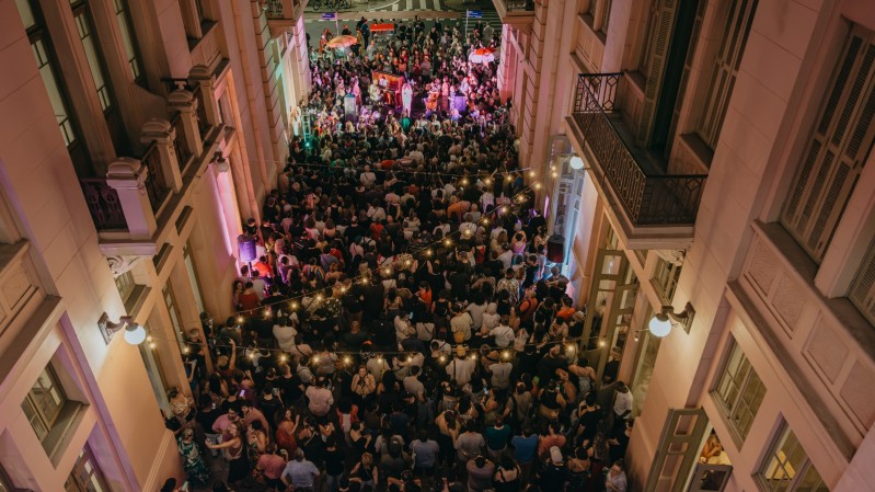 Vista aérea de um corredor estreito entre prédios históricos, lotado de pessoas assistindo a um show noturno ao ar livre, com iluminação colorida e cordões de luzes suspensas.