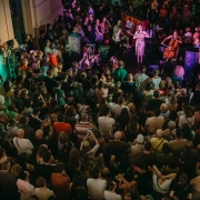 Multidão reunida diante de um palco pequeno em área externa, assistindo a uma cantora acompanhada por músicos, sob luzes coloridas e clima festivo noturno.