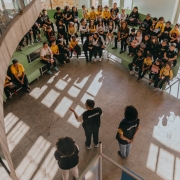 Grupo de crianças e adolescentes sentados em arquibancadas internas, ouvindo três mediadores uniformizados que conduzem uma atividade em um espaço iluminado pela luz do sol.