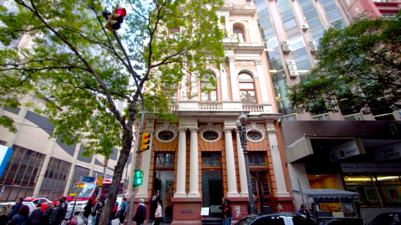 A imagem mostra, no centro, o edif&iacute;cio hist&oacute;rico de arquitetura cl&aacute;ssica do MuseCom com colunas cor&iacute;ntias, janelas ornamentadas e fachada em tons de bege e rosa claro. O pr&eacute;dio possui tr&ecirc;s andares vis&iacute;veis e est&aacute; espremido entre constru&ccedil;&otilde;es mais modernas, com fachadas de vidro e concreto.
&Agrave; frente do edif&iacute;cio, h&aacute; uma &aacute;rvore com galhos que se estendem por boa parte da imagem, parcialmente cobrindo a vis&atilde;o da constru&ccedil;&atilde;o. No canto superior esquerdo, h&aacute; um sem&aacute;foro com a luz vermelha acesa, e abaixo dele, uma luz de pedestre verde indicando que &eacute; seguro atravessar.
Na cal&ccedil;ada, v&aacute;rias pessoas caminham ou est&atilde;o paradas, e h&aacute; um carro preto estacionado em frente ao pr&eacute;dio hist&oacute;rico. &Agrave; direita, h&aacute; uma entrada de loja com toldo, e &agrave; esquerda, um caminh&atilde;o branco com detalhes vermelhos est&aacute; parcialmente vis&iacute;vel, provavelmente parado no tr&acirc;nsito.
