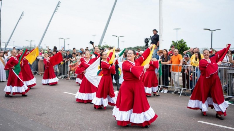 Mulheres dan&ccedil;ando na rua com vestidos vermelhos e bandeiras coloridas. Sorriem enquanto o p&uacute;blico assiste atr&aacute;s de notas. &Eacute; um momento alegre e cheio de movimento.
