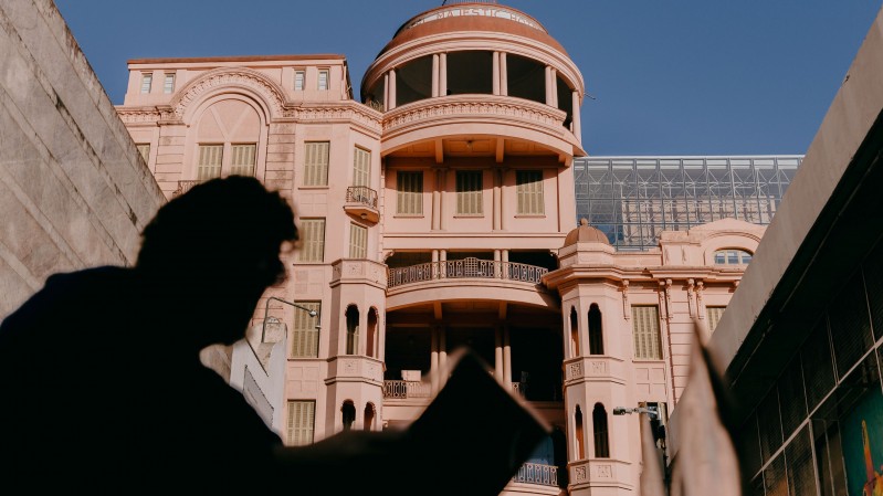 A imagem mostra a fachada da Casa de Cultura Mario Quintana de arquitetura imponente e detalhada, pintado em um tom rosado, com colunas e varandas decorativas. Em primeiro plano, h&aacute; a silhueta sombreada de uma pessoa lendo ou observando algo, criando um contraste marcante com a luz intensa que ilumina o pr&eacute;dio ao fundo. A sombra da pessoa parece estar em um beco ou corredor entre duas constru&ccedil;&otilde;es, direcionando o olhar para o edif&iacute;cio central da imagem. 