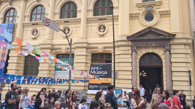 A imagem mostra a fachada do prédio histórico da Biblioteca Pública do Estado (BPE). A construção é de tom bege, com colunas, janelas altas e detalhes ornamentais. Na frente do edifício, há um evento acontecendo na rua: muitas pessoas caminham, conversam e observam barracas ou atividades. Bandeirinhas coloridas estão estendidas de um lado ao outro, criando um clima festivo. Também se veem cartazes e placas indicando programações culturais, incluindo um banner com o texto “BPE+ Música”. O ambiente é movimentado, com atmosfera de festival ou feira cultural ao ar livre, em frente à biblioteca pública.