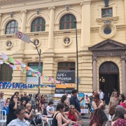 A imagem mostra a fachada do prédio histórico da Biblioteca Pública do Estado (BPE). A construção é de tom bege, com colunas, janelas altas e detalhes ornamentais. Na frente do edifício, há um evento acontecendo na rua: muitas pessoas caminham, conversam e observam barracas ou atividades. Bandeirinhas coloridas estão estendidas de um lado ao outro, criando um clima festivo. Também se veem cartazes e placas indicando programações culturais, incluindo um banner com o texto “BPE+ Música”. O ambiente é movimentado, com atmosfera de festival ou feira cultural ao ar livre, em frente à biblioteca pública.