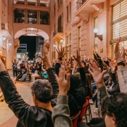 A imagem mostra um evento ao ar livre realizado em um corredor urbano entre prédios antigos, iluminado por luzes quentes, da Casa de Cultura Mario Quintana. Um grande grupo de pessoas está sentado e de pé, todas com os braços levantados, criando um clima de entusiasmo e participação coletiva. À esquerda, há um palco onde uma pessoa se apresenta ou conduz a atividade. Ao fundo, vê-se uma passagem em arco que revela a rua e a cidade à noite.