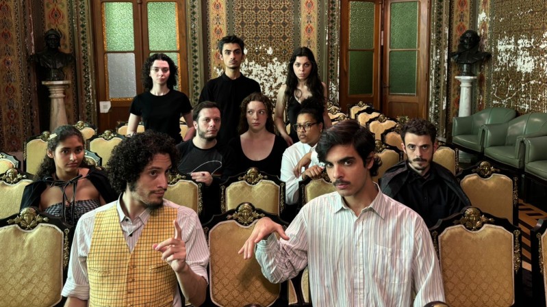 Fotografia de um grupo teatral, posando no Sal&atilde;o Mourisco da Biblioteca P&uacute;blica do Estado. &Eacute; uma sala ornamentada, com paredes decoradas com padr&otilde;es geom&eacute;tricos, muitos deles um pouco desgastados. H&aacute; dois bustos met&aacute;licos ao fundo duas grandes portas de madeira com vidro fosco. O ambiente inclui uma lumin&aacute;ria pendente central e poltronas verdes &agrave; direita. O grupo est&aacute; disposto em fileiras de cadeiras estofadas. S&atilde;o dez pessoas: cinco mulheres e cinco homens. Ao fundo, tr&ecirc;s atores est&atilde;o em p&eacute; e &agrave; sua frente tr&ecirc;s est&atilde;o sentados. Nas fileiras de cadeiras &agrave; frente est&atilde;o os demais quatro atores.  



