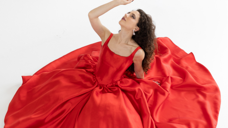 A imagem mostra a cantora Laura Dalm&aacute;s, uma mulher sentada sobre um amplo vestido vermelho de tecido brilhante, que se expande ao seu redor formando um c&iacute;rculo  volumoso e elegante. O vestido tem al&ccedil;as largas e caimento sofisticado. Ela est&aacute; com o corpo levemente inclinado para tr&aacute;s e o bra&ccedil;o direito levantado, como se estivesse alcan&ccedil;ando algo acima dela. Seu cabelo &eacute; longo, cacheado e solto, caindo sobre os ombros. O fundo &eacute; totalmente branco, destacando ainda mais o vermelho intenso do vestido e a pose expressiva da mulher.