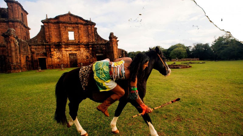 Na imagem, v&ecirc;-se uma pessoa montada a cavalo em um amplo campo gramado. O cavalo &eacute; escuro, com patas claras, e est&aacute; em movimento. A pessoa usa roupas coloridas e tradicionais, com franjas e detalhes chamativos, e parece inclinada para frente sobre o animal, segurando um objeto alongado, semelhante a uma lan&ccedil;a ou bast&atilde;o decorado.
Ao fundo, h&aacute; uma constru&ccedil;&atilde;o antiga de pedra avermelhada, parcialmente em ru&iacute;nas, com uma fachada imponente e janelas abertas, sugerindo um antigo templo ou edif&iacute;cio hist&oacute;rico. 