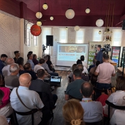 Sala com diversas pessoas sentadas, o Secretário da Cultura aparece de pé, ohando para uma tela de projeção com detalhes de um dos eventos.